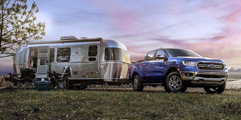 Brand new blue 2023 Ford Ranger pulling a travel trailer parked on a grassy lot with a lake in the background.